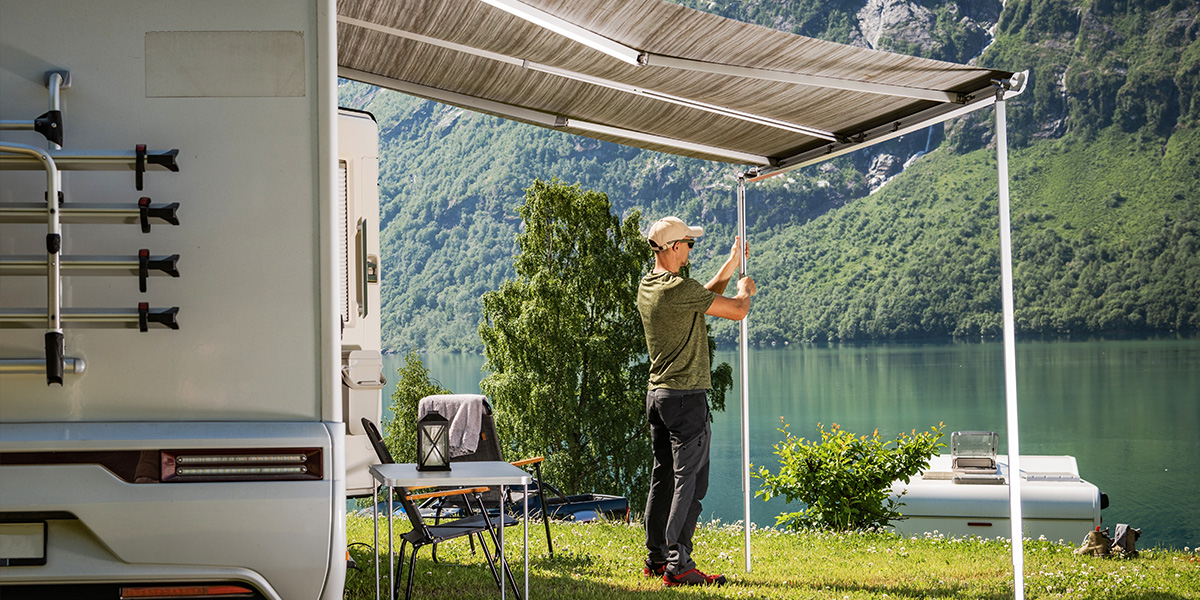 Man setting up an RV awning beside a scenic lake and mountains
