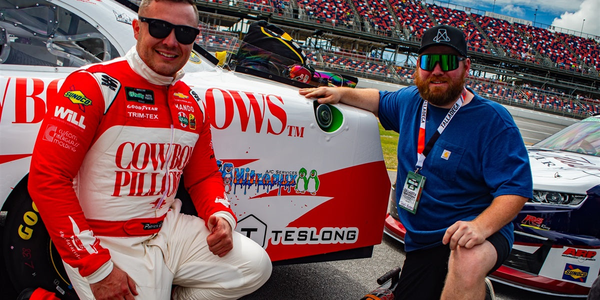 Race car driver and crew member pose beside a sponsored stock car at a racing track
