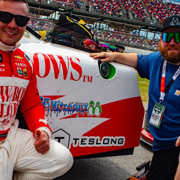 Race car driver and crew member pose beside a sponsored stock car at a racing track