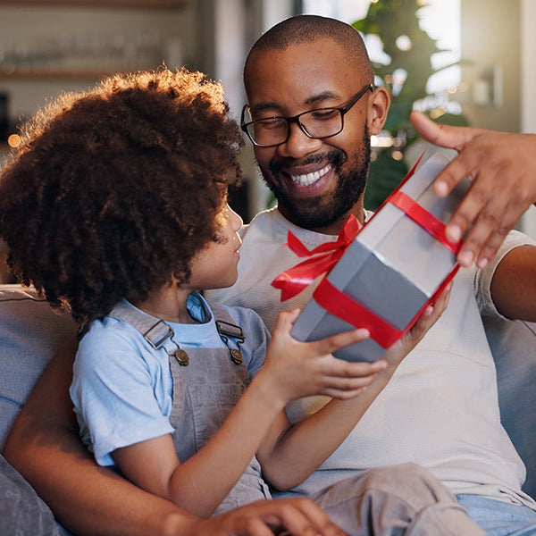 Child gives a wrapped Father’s Day gift to an adult while sitting together on a sofa at home