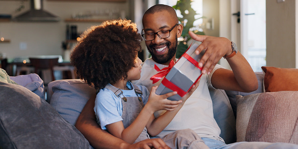 Child gives a wrapped Father’s Day gift to an adult while sitting together on a sofa at home