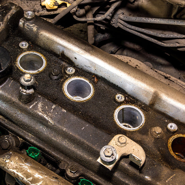 Dirty vehicle engine with the valve cover removed, exposing open spark plug wells and internal components